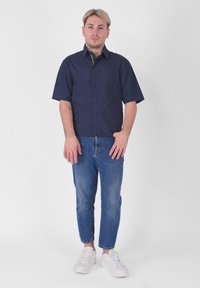 Camicetta blu a maniche corte, con tessuto leggero, abbottonatura frontale, abbinata a jeans blu a gamba dritta e scarpe da ginnastica bianche, in piedi su uno sfondo bianco.