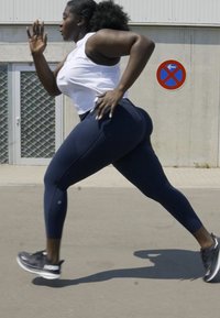 Mujer atlética negra corriendo con una camiseta sin mangas blanca y leggings azul marino, usando zapatillas de correr negras, con una superficie texturizada de fondo.