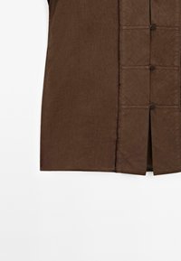 Camicia marrone con chiusura frontale con bottoni. Presenta un tessuto strutturato, design senza colletto e dettagli orizzontali cuciti lungo l'apertura.