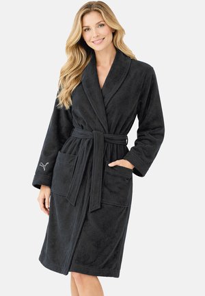 Lächelnde Frau, die einen schwarzen knielangen Mantel mit Gürtel und Taschen trägt, steht vor einem neutralen Hintergrund mit einer Hand in der Tasche.