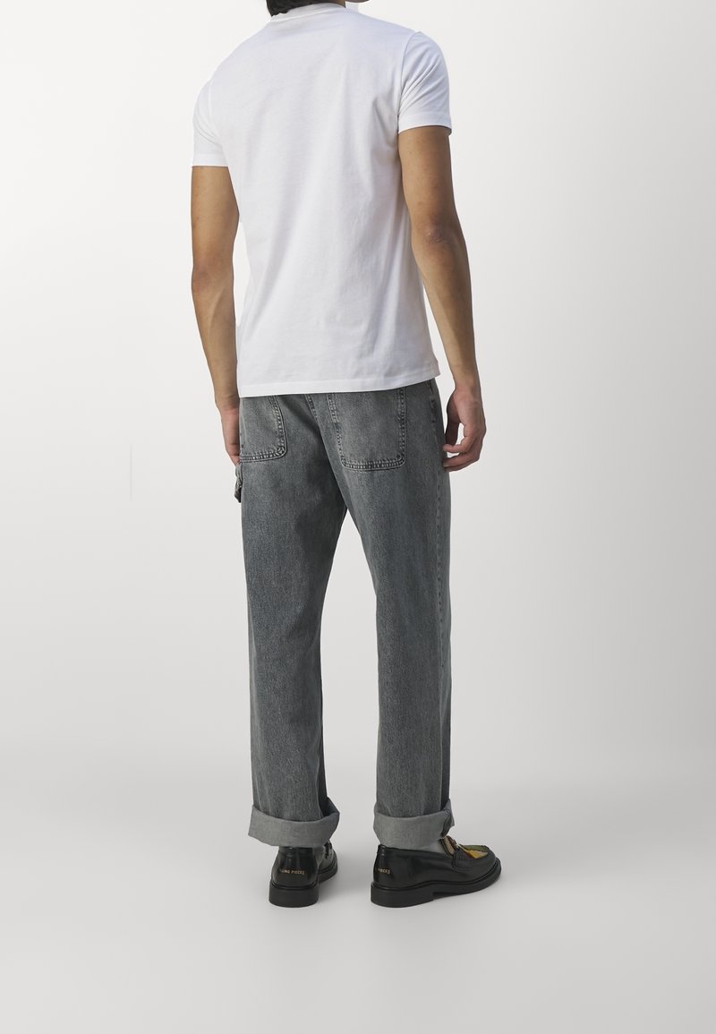White short-sleeve t-shirt paired with loose-fitting, rolled-up dark gray jeans. Model wears black shoes with yellow accents.