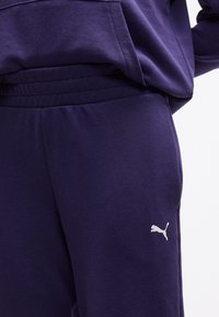 Primo piano di una persona che indossa pantaloni della tuta Puma color viola scuro e felpa abbinata con cintura elastica, su sfondo bianco.