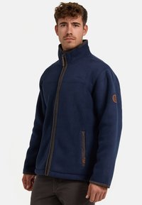 Marineblaue Fleecejacke mit hohem Kragen, kontrastierenden grauen Reißverschlüssen und einem Emblem am rechten Ärmel. Schlichtes Design mit seitlichen Taschen.