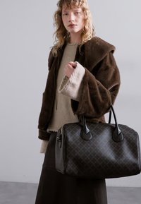 Donkerbruine nep-vachtjas over een beige trui, met een zwart geruit sporttas met ronde handvatten en ritssluiting.