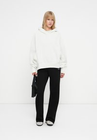 Felpa bianca oversize con cappuccio regolabile, abbinata a pantaloni nero svasati e sneakers grigie. Una borsa nera è tenuta in una mano.