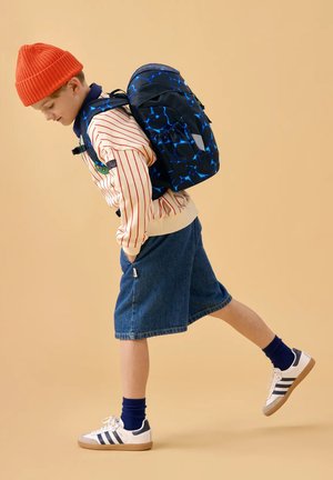 Junge mit orangener Mütze, gestreiftem Pullover, Jeansshorts und Turnschuhen trägt einen blauen Rucksack mit Muster und streckt das linke Bein aus, als würde er einen Schritt nach vorne machen.