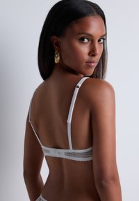 Soutien-gorge en dentelle blanche avec bretelles réglables et un design texturé, mettant en avant des motifs complexes le long de la bande et des détails délicats.