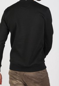Sudadera negra con cuello redondo, puños y dobladillo acanalados. Llevada con pantalones marrón claro. Textura suave con detalles mínimos.