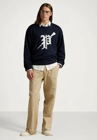 Ung man med glasögon som bär en marinblå tröja med vit "P" och pil-mönster, beige byxor, vit skjorta och vita sneakers, stående inomhus.