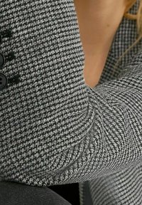 Primo piano di una persona che indossa una giacca con motivo pied de poule nero e bianco con bottoni neri, mostrando parte del collo e del braccio.