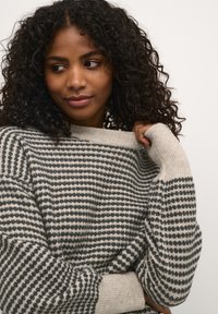 Pull en tricot avec un motif géométrique en gris et beige, doté d'un col large et de poignets côtelés. Matière en laine texturée.