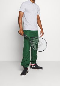 Man som bär grå polo, gröna joggingsbyxor och svarta Nike-sneakers, håller en svart och vit tennisracket framför vit bakgrund.