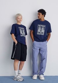T-shirts gráficos em azul-marinho apresentam uma impressão de pegada de sapato, combinados com calções pretos e calças azuis claras. Ténis texturizados completam os conjuntos.