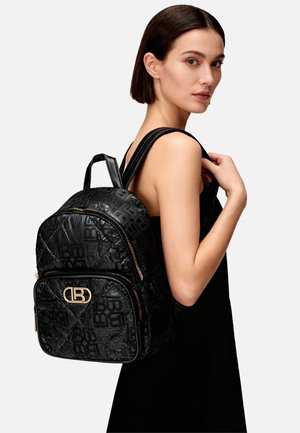 Zaino nero trapuntato con logo in rilievo, dettagli in metallo dorato, doppia cerniera e spallacci regolabili, indossato sulla spalla da una modella.