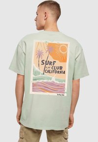 Jasnozielona bawełniana koszulka z retro grafiką na plecach, przedstawiającą palmy, fale oraz napis „Surf Club California.”