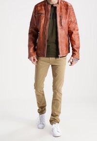 Brun läderjacka med dragkedja fram, olivfärgad t-shirt, beige byxor och vita sneakers. Tatuering synlig på handleden.