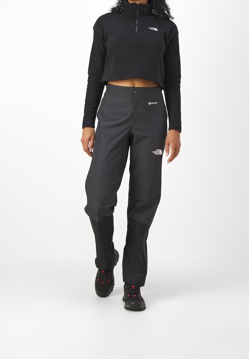 Femme portant un haut noir court à manches longues et un pantalon outdoor Gore-Tex noir avec des chaussures de randonnée, debout sur fond blanc.