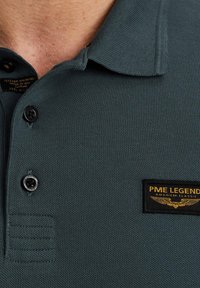 Tamnozelena polo majica s teksturiranim materijalom, s tri gumba na zapinjanju, crno-zlatnim logom "PME Legend" i rebrastim ovratnikom.