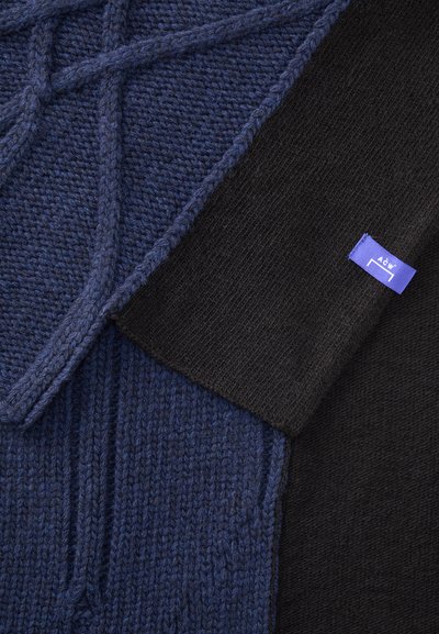 Close-up of blue cable-knit sweater with black sleeve featuring a small purple fabric tag labeled "A C W" on the cuff.