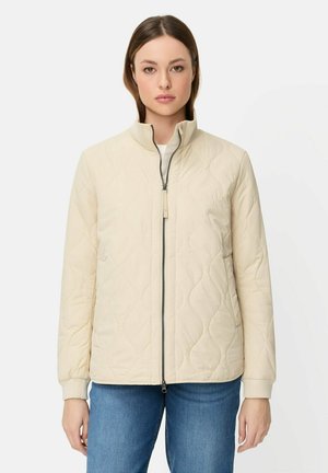 Kvinde iført en cremefarvet quiltet jakke med en lynlås foran, stående mod en ensartet hvid baggrund, kombineret med blå jeans.