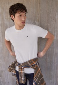 Camiseta blanca de manga corta con un pequeño logotipo negro en el pecho, combinada con una camisa de cuadros marrón, naranja y azul atada a la cintura.