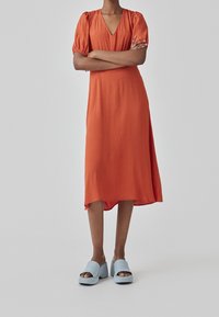 Robe midi orange avec manches bouffantes et col en V, présentant un ourlet légèrement asymétrique. Associée à des sandales à plateforme bleu clair.