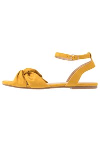 Keltainen mokkasiinisandaali, jossa on solmittu muotoilu pääpannassa ja nilkkapanta hopeaväljällä. Littee pohja, avokärkinen tyyli.