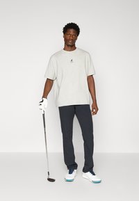 Szürke pamut póló logóval, fekete nadrág és fehér golfcipő kék részletekkel. Golf ütőt tart, semleges háttér előtt áll.