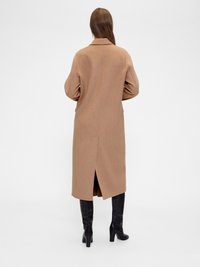 Long manteau couleur chameau avec un col classique, des épaules tombantes et une fente arrière. Porté avec des bottes noires ajustées montant jusqu'au genou. Tissu lisse.