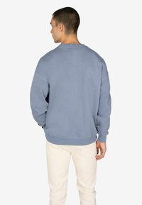 Felpa in cotone blu con maniche lunghe e scollo rotondo. Presenta polsini e orlo a coste, con una vestibilità rilassata. Indossata sopra pantaloni color crema.