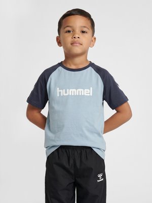 Młody chłopiec stoi ze splecionymi rękami za plecami, ubrany w niebieski t-shirt hummel i czarne szorty hummel na jednolitym tle.