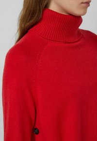 Czerwony sweter z golfem wykonany z dzianiny, z prążkowanym kołnierzem i guzikami po bokach. Gładka tekstura z dopasowanym krojem.