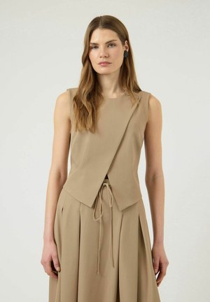Femme aux longs cheveux portant un top beige asymétrique sans manches et une jupe plissée assortie avec une taille à cordon, debout devant un fond uni.