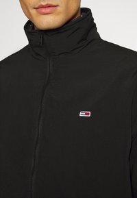 Black jacket with high collar, front zipper, and a small embroidered logo featuring blue, red, and white colors on the chest. Lightweight material.