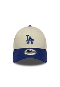 Casquette de baseball beige avec une visière bleue, présentant un logo "LA" brodé. Sangle ajustable à l'arrière et motif de tissu texturé sur la visière.