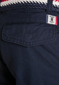 Námorně modré látkové šortky s tkaným pasem, který má červené a bílé akcenty; zadní kapsa s jemným prošíváním a značkovou visačkou.