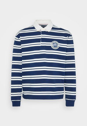 Tums zilas un baltas svītrots garām piedurknēm polo krekls ar baltu apkakli un apļa "NYC Columbus Avenue" logotipu uz krūšu.