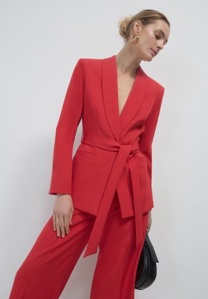 Roter taillierter Blazer mit tiefem V-Ausschnitt und gebundenem Taillengürtel, kombiniert mit roten weit geschnittenen Hosen. Strukturierter Stoff, minimalistisches Design und keine sichtbaren Verschlüsse.