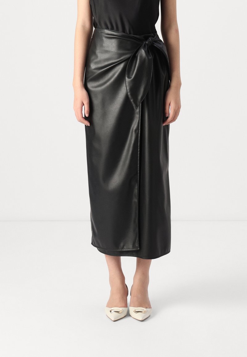 Femme portant une jupe midi portefeuille en cuir synthétique noir nouée à la taille et des talons blancs à bout pointu avec des détails de boucle dorée.