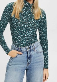 Bodysuit a maniche lunghe con una base verde scuro e modelli floreali bianchi e celesti sparsi, abbinato a jeans in denim azzurro a vita alta.