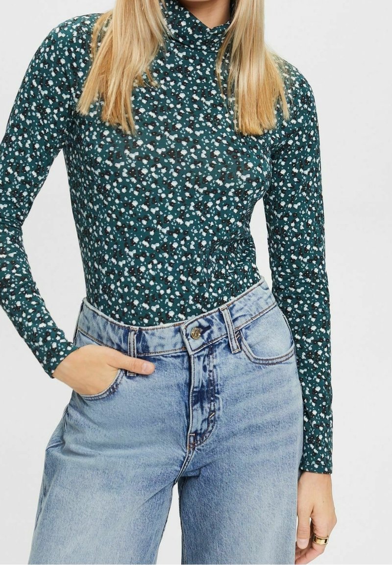 Bodysuit a maniche lunghe con una base verde scuro e modelli floreali bianchi e celesti sparsi, abbinato a jeans in denim azzurro a vita alta.