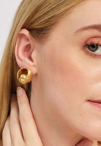 Zeltainas auskari ar skulpturālu dizainu, kurā ir noapaļota doba daļa un centrālais bumbulis, radot vizuālu dziļumu un tekstūru.