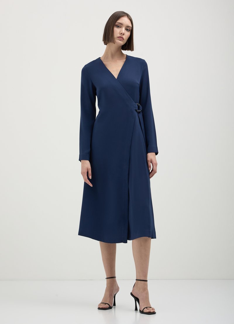 Robe portefeuille bleu marine à manches longues, avec un détail de boucle sur le côté. Fabriquée en tissu lisse, elle tombe jusqu'au milieu du mollet, complétée par des escarpins noirs.