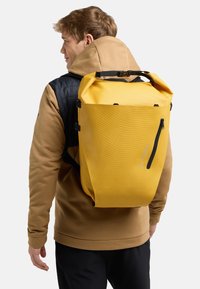 Mochila impermeable amarilla con una superficie texturizada, correas ajustables y un bolsillo con cremallera lateral, llevada en la espalda de una persona.