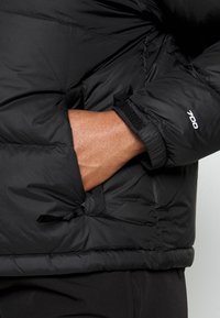 Mano insertada en el bolsillo lateral de una chaqueta acolchada negra con "700" impreso en la manga cerca de un puño de velcro.