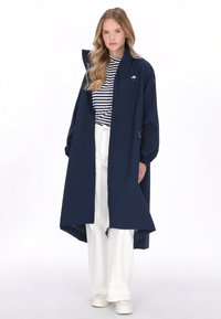 Tengerészkék kapucnis esőkabát laza szabással, könnyű anyagból, nagy első zsebekkel és rugalmas mandzsettákkal, csíkos ing és fehér nadrág felett viselve.