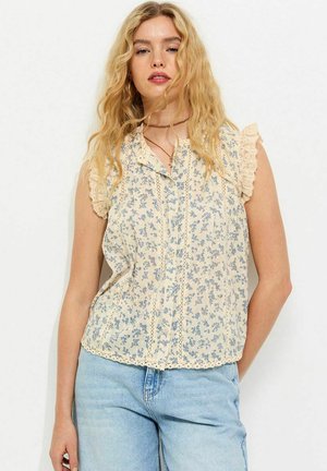 Młoda kobieta o falowanych blond włosach, ubrana w bezrękawową kremową bluzkę z niebieskim kwiatowym wzorem oraz jasnoniebieskie dżinsy z wysokim stanem.