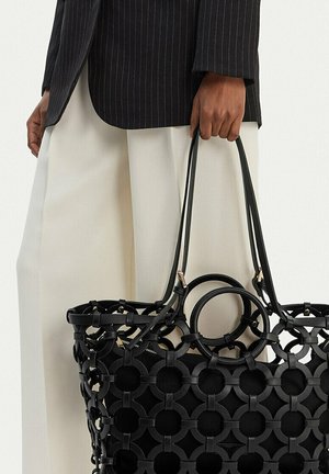 Mano sosteniendo un gran bolso tote negro con un patrón de recortes circulares, vistiendo una chaqueta negra de rayas y pantalones anchos color crema.