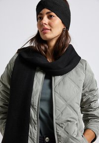 Diadema y bufanda de punto negras, usadas con una chaqueta gris acolchada y una blusa color teal. La bufanda cae de manera casual alrededor del cuello.