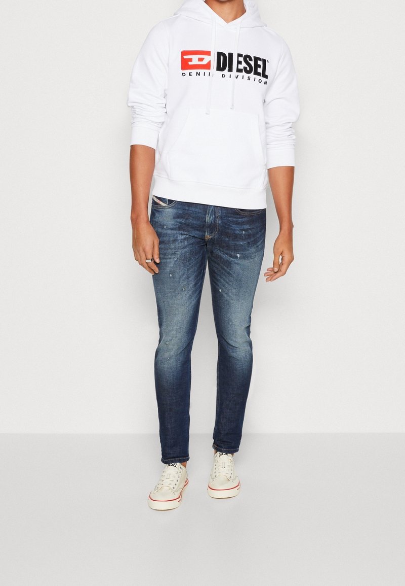 Personne portant un sweat à capuche blanc Diesel avec logo, jean slim bleu délavé et baskets blanches avec bordure rouge, debout sur un fond uni.